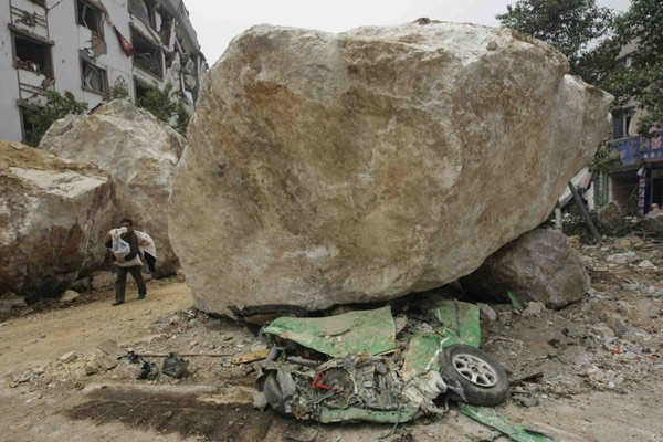 5月16日，四川北川县城，一位灾民带着行李撤离危险地区，道路旁边，一块巨大的滚石把一辆汽车砸扁。