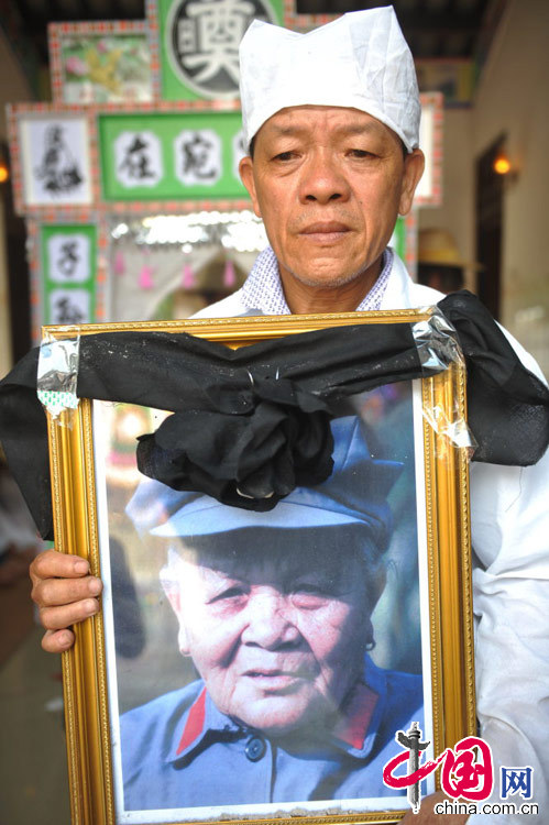 2014年4月20日，海南琼海，卢业香的儿子翁祚雄手捧母亲遗像走出大门。当日，最后娘子军卢业香的葬礼在其家乡海南省琼海市中原镇仙村村隆重举行。中央芭蕾舞团、琼海市委市政府等单位和部门送了花圈。