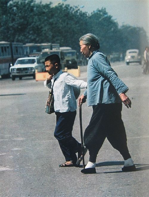На снимке: старушка с перебинтованными ногами переходит улицу, держа под руку школьника.