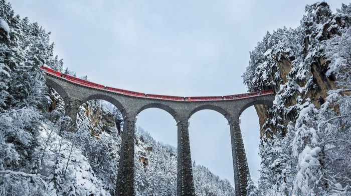 Сказочный пейзаж: красный поезд проходит через столетний мост в снегу