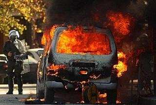 A car burns in front of riot police during a massive demonstration near the main police station in the northern Greek city of Thessaloniki. [Sakis Mitrolidis/AFP]