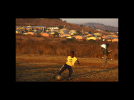 A local child plays soccer at a dirt field in Soweto, Johannesburg June 7, 2010, as a man walks towards homes carrying a tray of eggs and a sack of flour. [China Daily/Agencies]