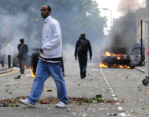 A man walks past a lit car on a street in Hackney, east London, Britain, Aug. 8, 2011. Rioting starting late Saturday appeared spreading across London on Monday, with vandalising, arson and looting taking place in various London communities. [Zeng Yi/Xinhua]