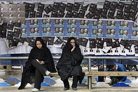 Two Iranian women are waiting for the bus. Election posters can be seen on the wall behind them. 