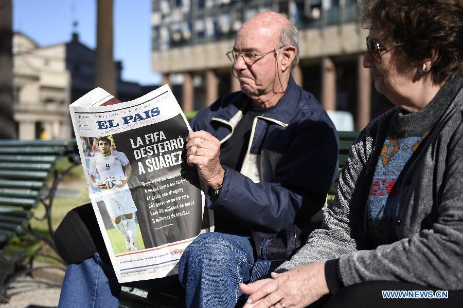 A woman （R）looks at a newspaper with the image of Uruguayan football player Luis Suarez, in Montevideo, capital of Uruguay, on June 27, 2014. Luis Suarez was banned by FIFA for four months from any football-related activity on June 26 for biting Italy&apos;s player Giorgio Chellini in a Group D match between Italy and Uruguay on June 24.