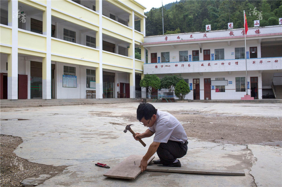 Xiong Chaogui makes a board on which he plans to write “Be careful” to put it on the low wall at the gate for the safety of the students. As the conditions at the school are fairly poor, it’s hard to attract other teachers. Xiong does every small work by himself. [Photo/qq.com]
